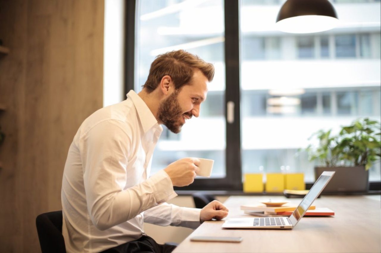 man-at-desk-holding-cup-of-coffee-looking-pleased-at notebook-coffee-cup-daytime-925786